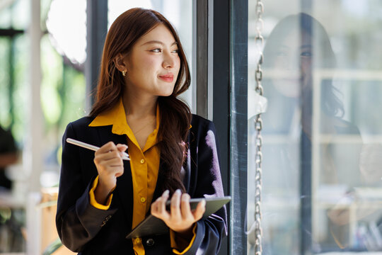 Medium shot of smiling asian businesswoman taking notes in notebook while working or studying at contemporary office center, copy space
