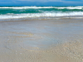 Low-angle beach view with rippled wet sand, tiny bubbles, blurred teal waves with foamy crests, and a clear blue sky with soft clouds, creating a dreamy, tranquil mood.