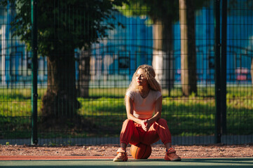 Young woman sitting on a basketball with sunlight on her face at outdoor court, wearing coral pants and beige top, resting during game in urban park.