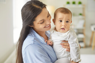 A smiling mother holds her curious baby toward the camera in a bright bedroom, making a warm portrait that shows close bond and gentle interaction. Mother holds her curious infant to camera.