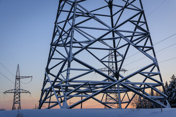Snow-Covered High Voltage Power Pylons at Sunset in Winter Landscape, Industrial Infrastructure Against Cold Blue Sky and Orange Horizon, Energy Grid in Nature