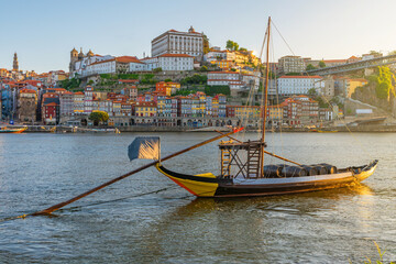 Traditional rabelo boat on the Douro River with colorful Ribeira district and historic buildings in Porto city, Portugal. Hillside cityscape with Clerigos Tower and Dom Luis Bridge at sunrise