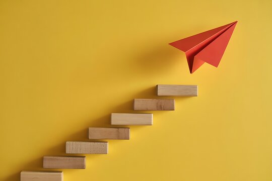 A red paper airplane ascends a staircase of wooden blocks against a yellow background, symbolizing progress, growth, and the achievement of goals through incremental steps