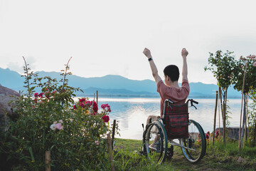 Rear view of young man with disability in wheelchair on the nature at sunset, He is arms raised with confident, Concept of healthcare for person with disabilities,rehabilitation with natural therapy.