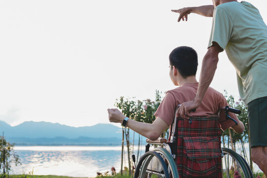 A young man with disability in wheelchair and his parent on the lake at sunset with background of the hill, Health care for the sick or person with disabilities, rehabilitation with natural therapy.