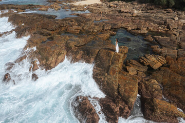 Woman in White Dress Standing on Rocky Shore with Crashing Waves