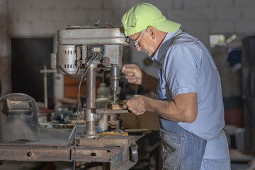Senior craftsman drilling metal workpiece using a drill press in workshop, demonstrating skill and experience