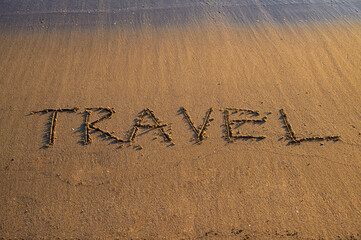 The word “TRAVEL” written on sandy beach near the shoreline, with gentle sea waves approaching.