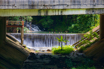 Bridge over a peaceful river with a waterfall surrounded by lush trees in Punyaban Waterfall, Ranong, Thailand.