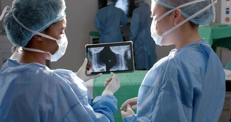Examining gowned surgeons holding tablet showing spinal x-rays around surgical table in OR