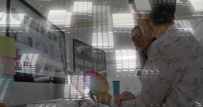 Designer in floral blouse examining monitors displaying designs at office desk, holding color wheel