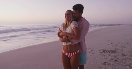 Embracing couple wearing beachwear and resort clothing on sandy beach at dusk with pastel sky