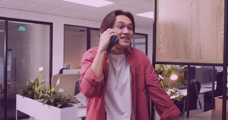 Leaning Asian man in white T-shirt, red shirt speaking on smartphone in office by string-lit plant