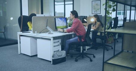 Working Asian male using monitor and laptop at open-plan office with divider screens and plants