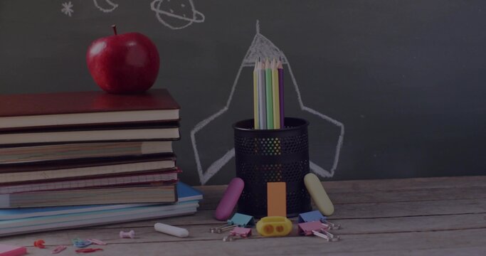 Holding colored pencils, pencil holder sitting in class with apple-topped textbooks and chalkboard
