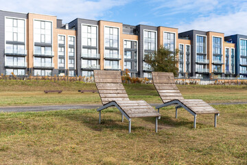 Two wooden lounge chairs in grassy area with modern apartment building in background
