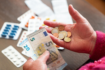 An elderly woman counting money against a background of scattered medicines. An elderly woman holding Euro banknotes and coins, the financial difficulties with the cost of medicines in old age