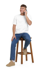 Handsome young man in stylish jeans talking on smartphone on stool against white background