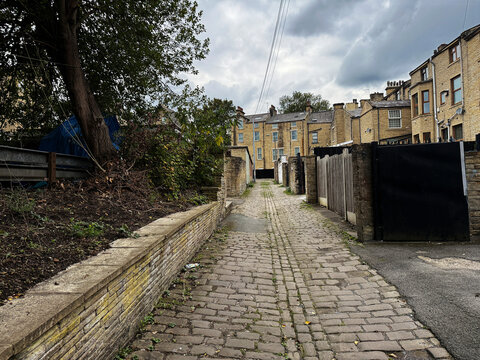 A Victorian cobblestone path meanders between weathered stone homes, dappled with shade from towering trees that lend the scene a hushed tranquility in Manningham, Bradford, UK