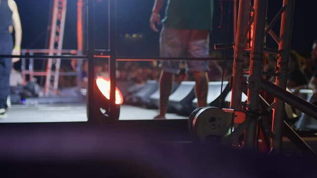 Side close-up of metal concert stage structure at night, with blurred performer and crowd in background. Captures live music energy and technical setup in a modern, atmospheric scene.