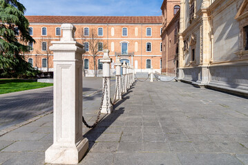 Alcalá de Henares, Spain - April 9, 2025: Outdoor plaza at the University of Alcalá, featuring stone bollards and chains, separating the main ornate stone building from a large, red-brick structure.