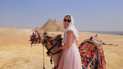 Woman enjoying a vacation in Egypt, posing with a decorated camel near the Pyramids of Giza under a clear blue sky.