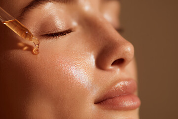Golden skincare cosmetic serum pouring and flowing from pipette on beautiful female woman beauty models face detail. Gentle natural moisturizing hydration and skin care routine.