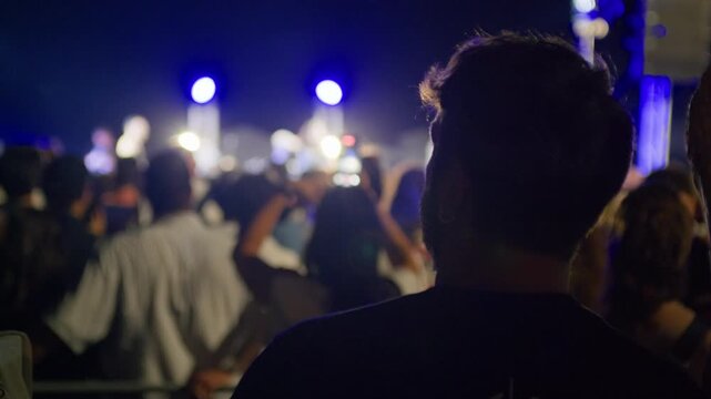 Rear view of a young man on his phone during a nighttime concert. The crowd and stage blur behind, highlighting digital distraction amid a live music experience.