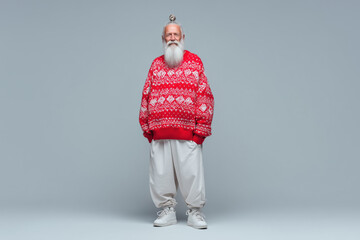 Senior Caucasian man standing and smiling with hands in pockets, wearing patterned sweater and loose pants, long beard and hair tied in top knot, posing against plain background