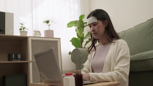 Working from home concept. Young Asian woman employee is symptoms sick and has a headache from the flu use fever cooling gel patch and sitting using laptop computer for working in living room at home.