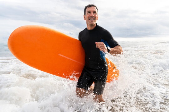 Middle aged sportive handsome man learning surfing and running in the waves holding board
