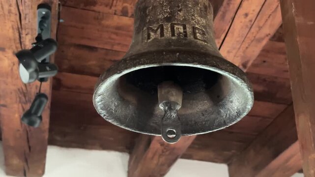 A close-up of a dark, old metal bell hangs from wooden beams inside a tower or belfry; the inscription "MRE" is faintly visible near the rim