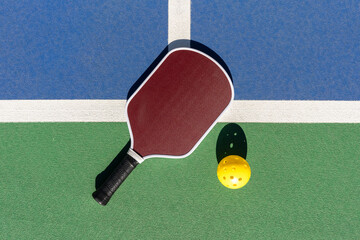 Top-down high-angle view of pickleball paddle and yellow ball on green and blue outdoor court surface