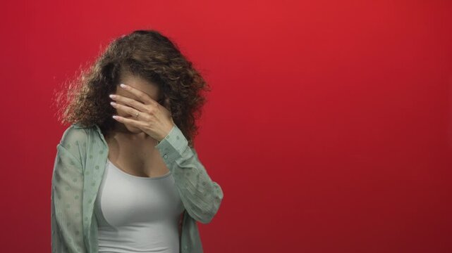 Woman with hand to forehead wearing white camisole and sheer green shirt in red studio; shy introspection quiet.