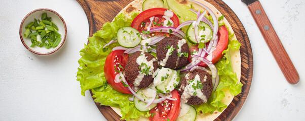 Preparing falafel wraps with crisp lettuce, sliced tomatoes, cucumbers, and red onions drizzled with tahini sauce, a vegetarian snack on a rustic cutting board with hummus