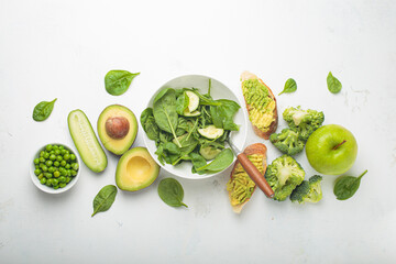 Fresh green salad bowl, avocado toasts, raw vegetables fruit white concrete background top view, diet, vegetarian food. Set of different green healthy products, clean eating nutrition