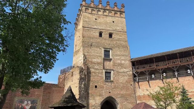 Tall medieval brick tower with crenellations and arched gate, next to a wooden defensive gallery and wall, framed by green trees under a clear blue sky