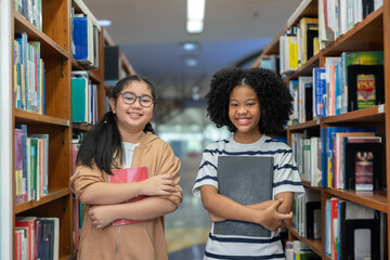 Happy multicultural student girls standing between library bookshelves, holding books and smiling confidently in educational school.