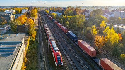Skalmierzyce railway station. Drone view of railway tracks with a passenger train and container freight train in autumn trees and urban surroundings.