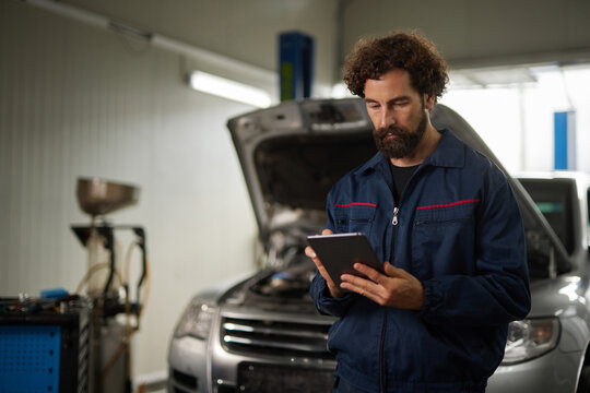 Car mechanic stands in a well-lit auto repair shop, focused on a tablet while inspecting a vehicle with an open hood for diagnostic purposes. - Powered by Adobe