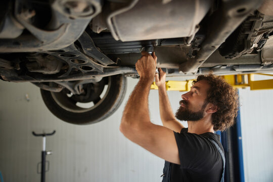 A skilled mechanic is working under a car in an auto repair shop during the afternoon. He is using tools to check and fix parts while ensuring everything runs smoothly. - Powered by Adobe