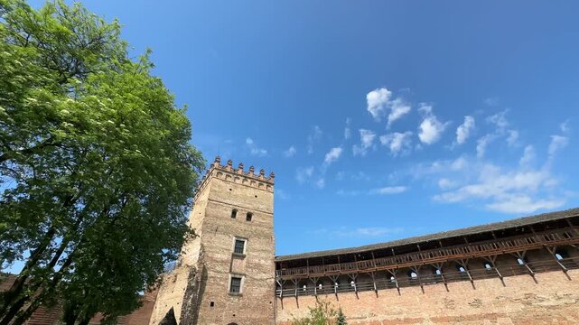 Low-angle view of a tall medieval brick tower with crenellations and an adjoining long curtain wall topped by an extensive wooden fighting gallery under a vibrant blue sky