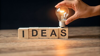 Illuminating ideas hand placing light bulb atop wooden blocks spelling ideas on a table, symbolizing creativity and innovation on a dark background