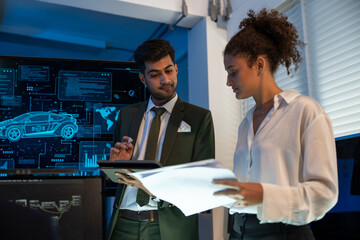 Two professionals analyzing and discussing documents with digital car interface in high-tech automotive innovation at dark modern office.