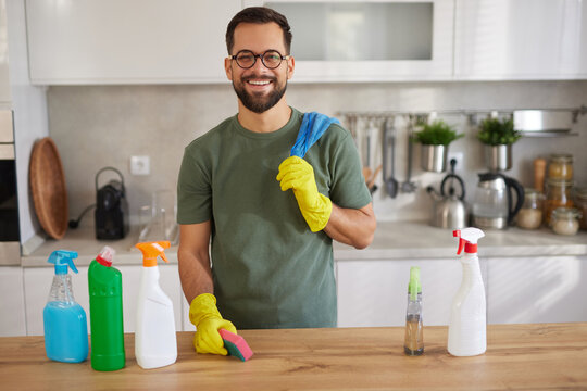 A cheerful man is preparing for house cleaning in a modern kitchen. He holds a sponge and stands among various cleaning supplies, ready to tackle the chores.