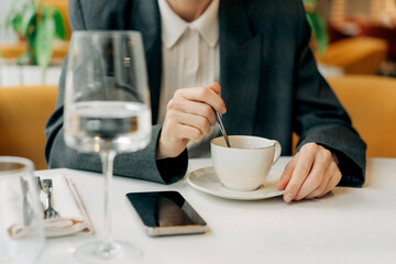 Close-up of an unrecognizable woman's hands placing sugar in coffee in a restaurant while her mobile phone lies nearby on the table.