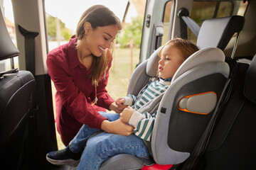 A mother is securing her young child in a car seat while smiling warmly. They are parked in a...