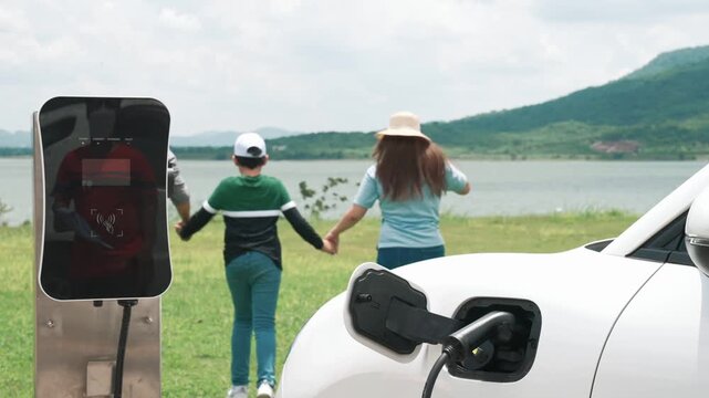 Concept of progressive happy family enjoying their time at wind farm with electric vehicle. Electric vehicle driven by clean renewable energy from wind turbine generator for charging station.