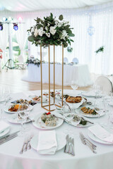 Close-up of a perfectly set wedding dinner table featuring a tall golden stand with a white and green floral arrangement, plates with appetizers, and sophisticated glassware