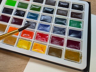 Watercolor paint palette with brush ready to use. Close-up of a colorful watercolor paint set with a brush, placed on a table next to blank white paper